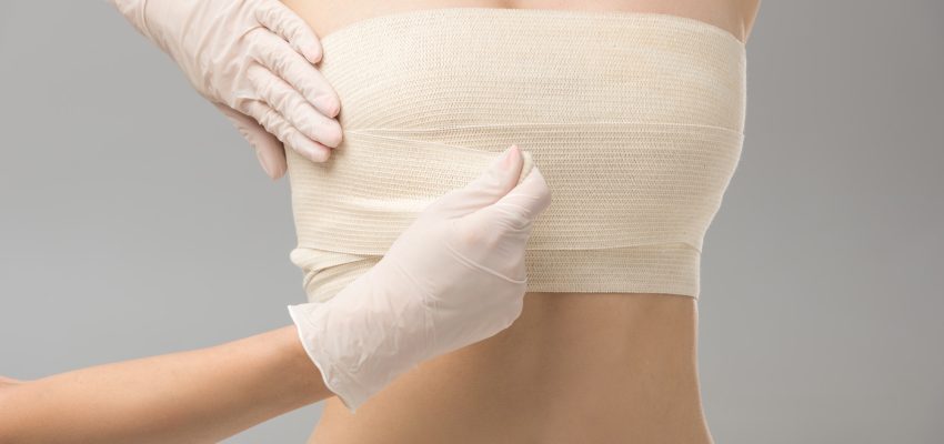 partial view of plastic surgeon in latex gloves and patient in breast bandage isolated on grey